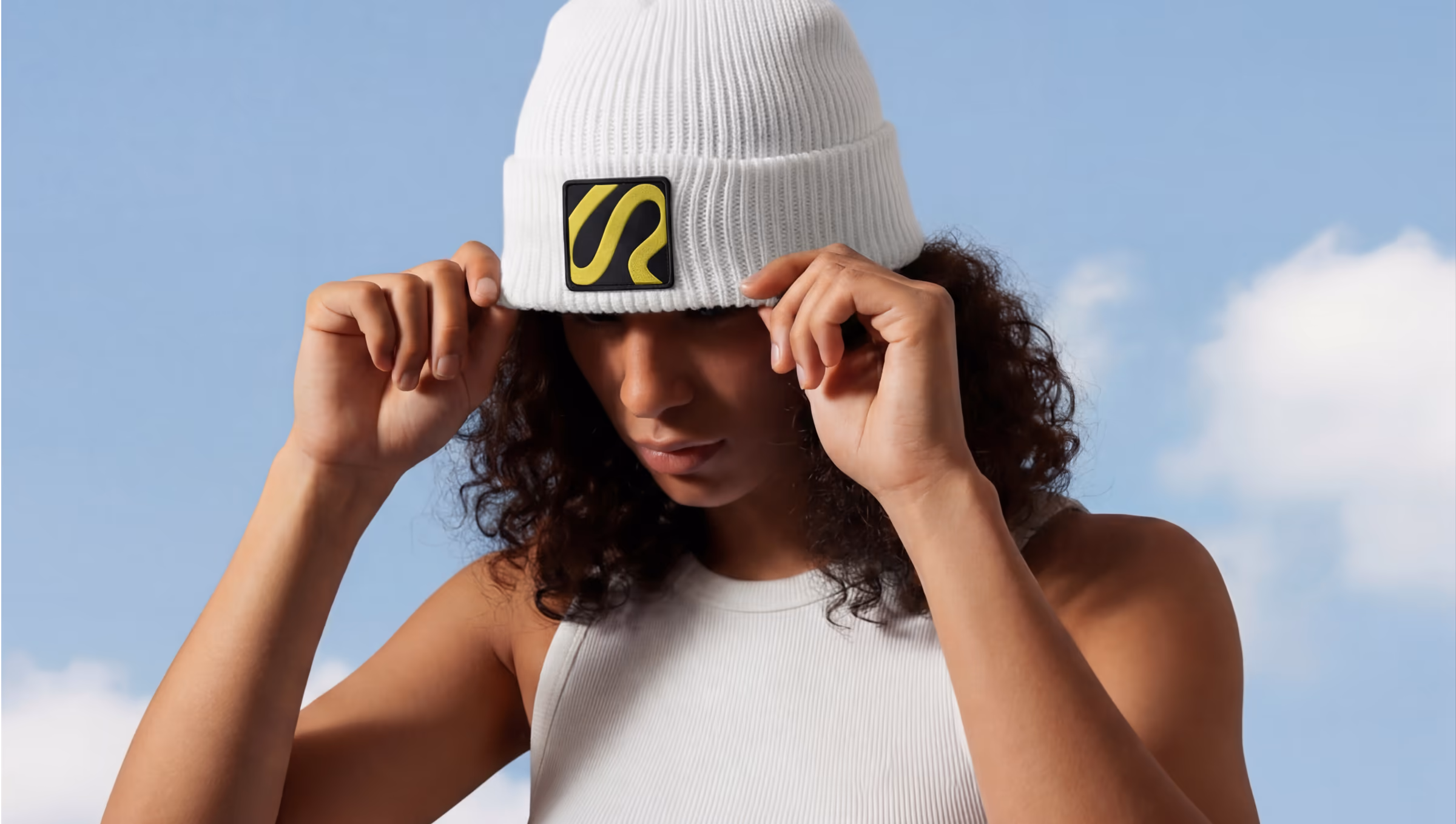 Woman adjusting a white beanie with a yellow and black Riverflow logo patch, against a blue sky