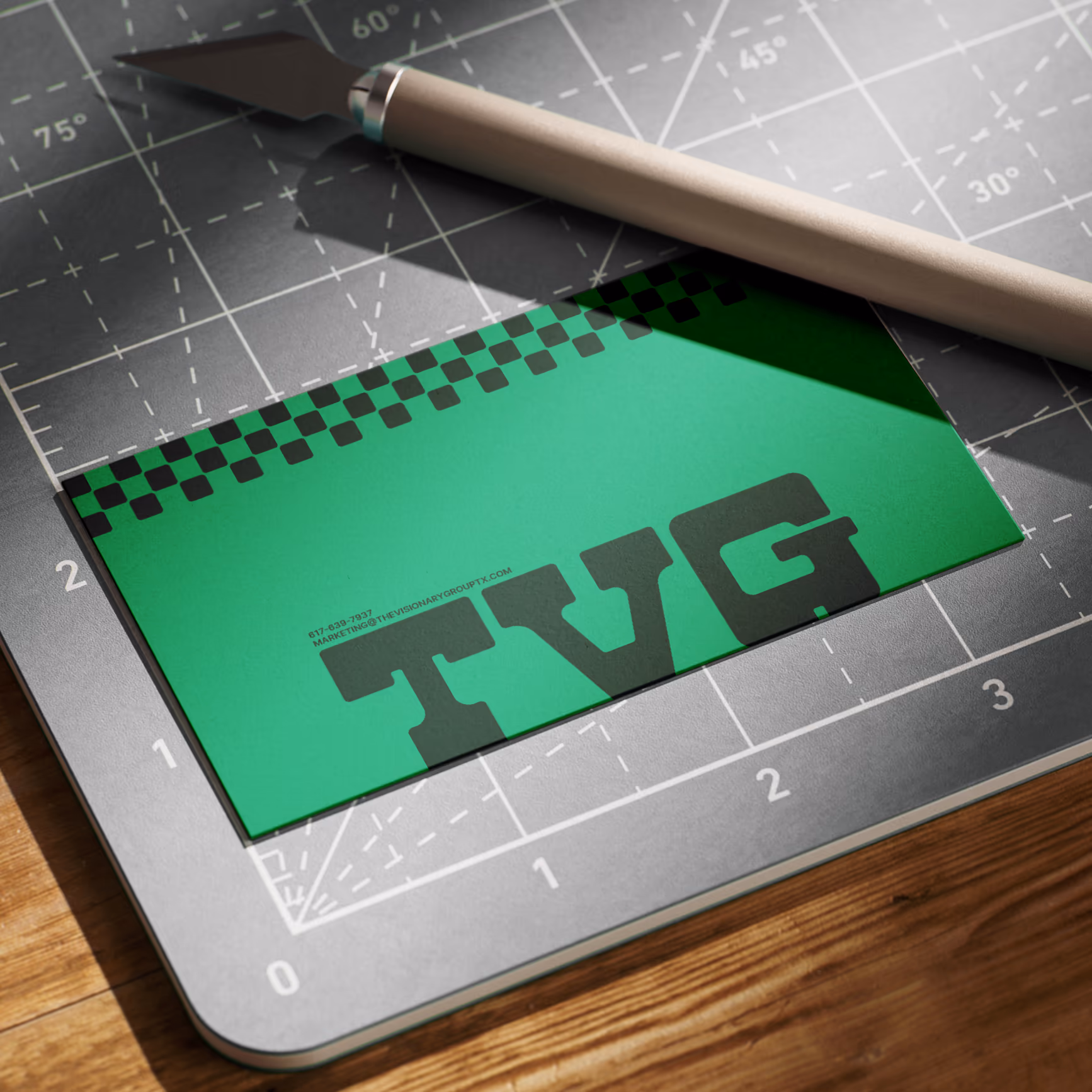 Green TVG business card with slab wordmark and checkered strip, on a cutting mat with a knife